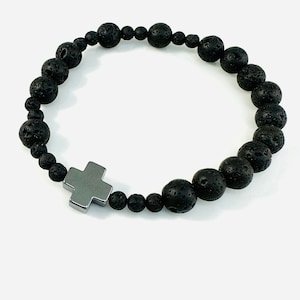 Könnte beinhalten: Ein schwarzes Perlenarmband mit einem silbernen Kreuzanhänger. Die Perlen sind aus Lavastein.