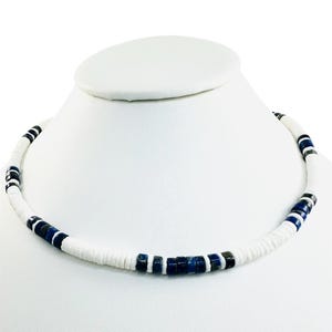Può includere: Una collana di perline bianche con sezioni alternate di perline bianche, blu scuro e nere. La collana è esposta su un collo di manichino bianco, evidenziando il suo design e il contrasto cromatico. Le perline sono cilindriche e uniformemente distanziate.