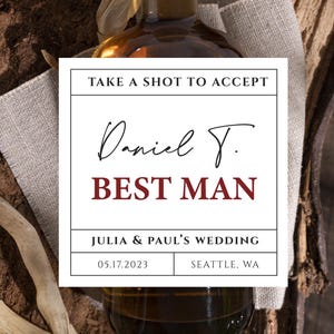 May include: A glass bottle with a wooden stopper, containing a dark amber liquid. The label reads "TAKE A SHOT TO ACCEPT" and "Daniel T. BEST MAN" for a wedding in Seattle, WA on 17.05.2023. The bottle is on a wooden surface.