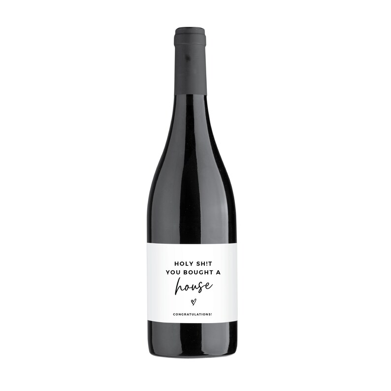 Puede incluir: Una botella de vino tinto con una etiqueta blanca que dice "Holy Shit You Bought A House" con un coraz&oacute;n y la palabra "Congratulations!"