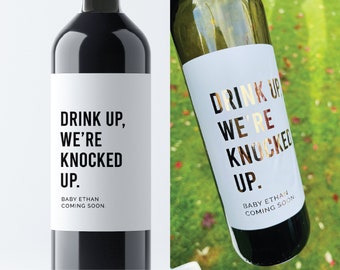 May include: Two wine bottles with white labels. The labels have black text that reads "Drink Up, We're Knocked Up. Baby Ethan Coming Soon."