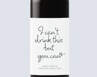 May include: A white wine bottle label with black text that reads "I can't drink this but you can" with a heart symbol. Below the text is "Baby Martin Arriving April 2023".