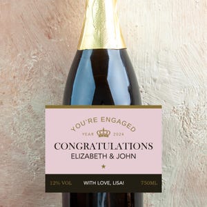 May include: A champagne bottle with a gold foil top and a pink label. The label reads "YOU'RE ENGAGED" with "CONGRATULATIONS ELIZABETH & JOHN" below. The label also includes the year 2024, a crown, and the text "12% VOL WITH LOVE, LISA! 750ML".