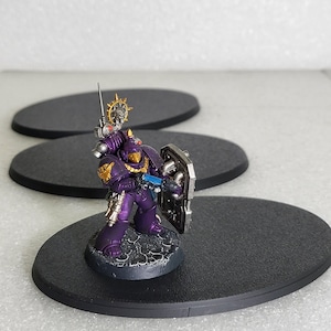 May include: A miniature figurine of a space marine in purple armor, holding a sword and shield. The figurine is standing on a black, oval base. Additional black bases are in the background. The space marine has gold and silver accents.