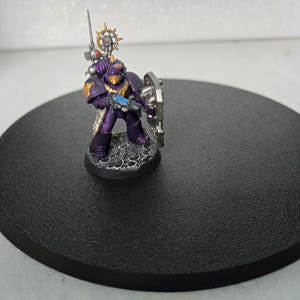 May include: A miniature figurine of a space marine in purple armor with gold accents, holding a weapon and shield. The figure stands on a textured base, set against a black circular platform. The miniature is detailed and painted.