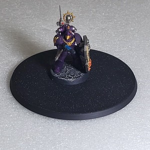 May include: A purple and gold painted miniature space marine figure holding a sword and shield, standing on a black circular base.