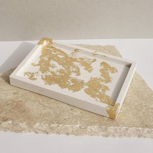 Könnte beinhalten: Ein weißes rechteckiges Tablett mit goldenen Blattverzierungen. Das Tablett ist aus Beton gefertigt und hat eine strukturierte Oberfläche.