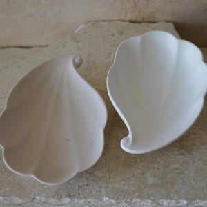 Könnte beinhalten: Zwei weiße, blattförmige Schalen aus Keramik. Die Schalen haben leicht unterschiedliche Formen und Größen. Die Schalen befinden sich auf einer hellbraunen Oberfläche.