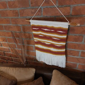 Può includere: Un arazzo da parete bianco, rosso e giallo tessuto con frange. L'arazzo è fatto di filo ed è attaccato a un bastone di legno.