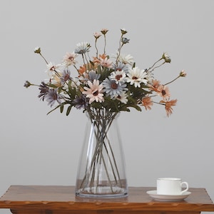May include: A bouquet of artificial daisies in shades of gray, brown, and white arranged in a clear glass vase on a wooden table. The vase is tall and slender with a rounded base.