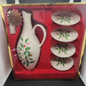 May include: A white ceramic pitcher with a green holly and red berry design. The pitcher is in a red box with four white ceramic plates with the same holly and berry design. The box has a gold border and the words "Great Gift" on a gold star.