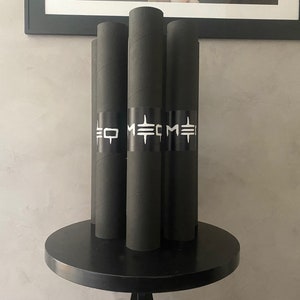 May include: Four black cylindrical tubes with white text and a black band around the middle, standing upright on a black round table. The text on the band reads "EO METME". The tubes are likely posters or prints.