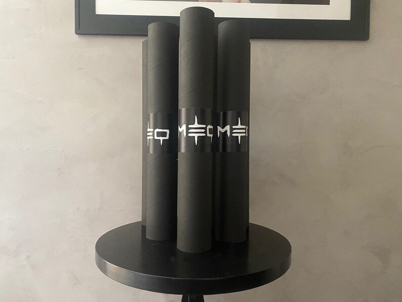 May include: Four black cylindrical tubes with white text and a black band around the middle, standing upright on a black round table. The text on the band reads "EO METME". The tubes are likely posters or prints.