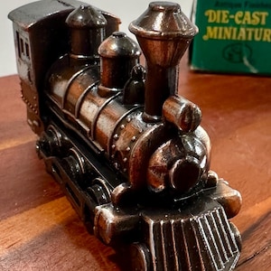 May include: A detailed die-cast miniature train engine with a metallic bronze finish. The model features intricate detailing, including a smokestack, cab, and wheels. The box next to the train reads "DIE-CAST MINIATURE".
