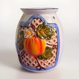 May include: White ceramic essential oil diffuser with a raised pumpkin design. The pumpkin is orange and yellow with green leaves and a white flower. The diffuser has a blue border and a lattice pattern background.