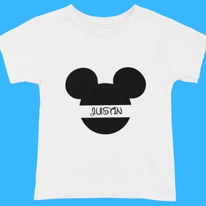 May include: White toddler t-shirt featuring a black Mickey Mouse silhouette with the name "JUSTIN" in a white horizontal band across the face. The shirt has short sleeves and a crew neck.