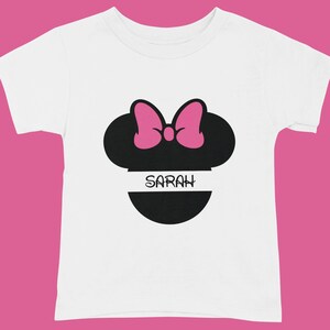 May include: White toddler t-shirt featuring a black silhouette of a mouse head with a pink bow. The name "SARAH" is printed in white on a black horizontal stripe below the mouse head design.