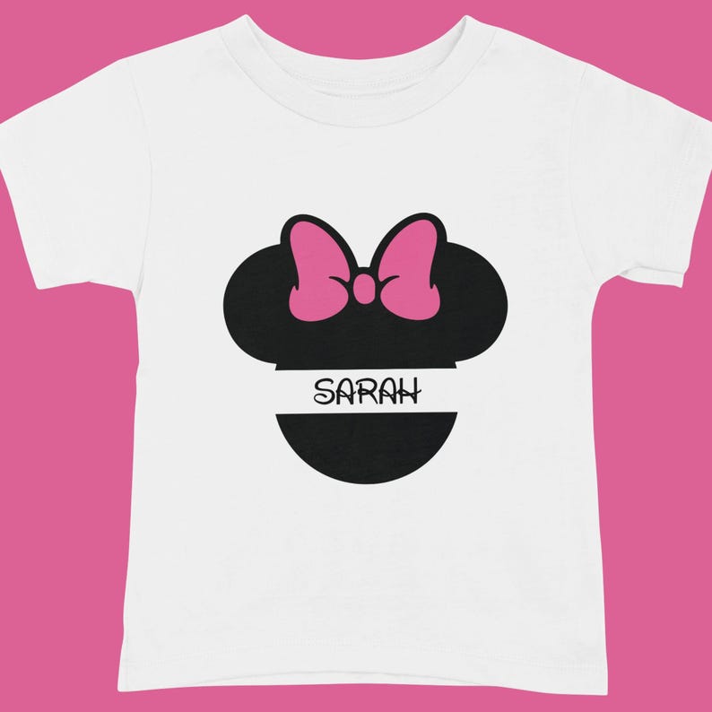 May include: White toddler t-shirt featuring a black silhouette of a mouse head with a pink bow. The name "SARAH" is printed in white on a black horizontal stripe below the mouse head design.