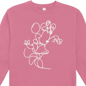 May include: A pink sweatshirt featuring a white line drawing of a cartoon character. The design is a continuous line, depicting the character's head, ears, and body. The sweatshirt has a crew neck and long sleeves.
