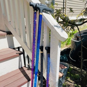 Puede incluir: Cuatro bastones de caminar con detalles decorativos de cristal. Los bastones son de color rosa, azul, negro y morado. Los bastones están apoyados en una barandilla de madera blanca.