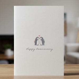 May include: A white card with a drawing of two penguins kissing with a small red heart above them. The text "Happy Anniversary" is written below the penguins.