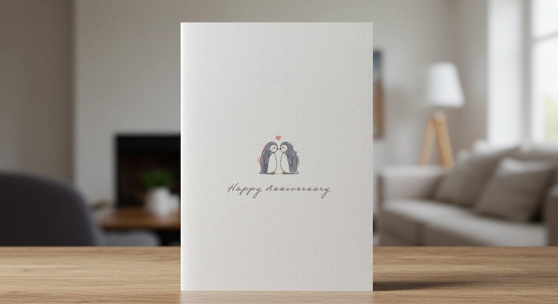 May include: A white card with a drawing of two penguins kissing with a small red heart above them. The text "Happy Anniversary" is written below the penguins.