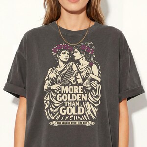 May include: Dark gray t-shirt featuring a graphic of two women with laurel wreaths, holding musical instruments. The text on the shirt reads "MORE GOLDEN THAN GOLD" and "THE LESBOS TOUR - 600 BCE". The shirt has a vintage, worn look.