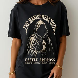 May include: Black t-shirt with a vintage-style design. It features a hooded figure holding a candle, with the text "THE BANISHMENT TOUR" above and "CASTLE ARDROSS" below. Roman numerals are also included in the design.