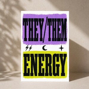 Op de afbeelding: Een rechthoekige kunstprint met de woorden "THEY/THEM" in zwarte blokletters op een paarse achtergrond, en "ENERGY" op een gele achtergrond. Symbolen van bliksemschichten, een maan en een ster zijn ook aanwezig.
