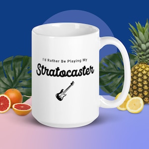 May include: White ceramic mug with the text "I'd Rather Be Playing My Stratocaster" and a black guitar illustration. The mug is surrounded by tropical fruit and foliage, including a pineapple, grapefruit, and lemon.