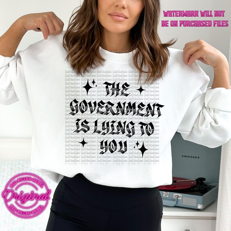 May include: White sweatshirt with the text "The Government Is Lying To You" in a gothic font. The sweatshirt has long sleeves and a crew neck. A pink and red logo is visible in the lower left corner.