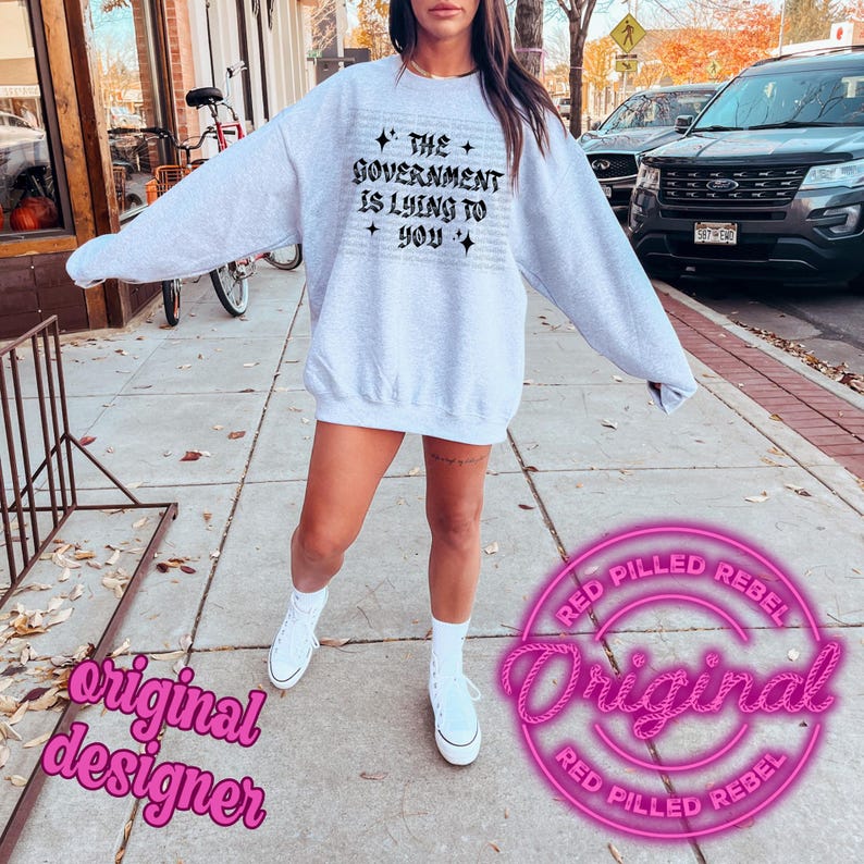 May include: A light gray sweatshirt with the text "THE GOVERNMENT IS LYING TO YOU" in a gothic font. The sweatshirt is worn with white sneakers and socks. A pink "Original" logo is visible.
