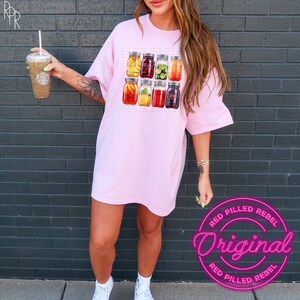 May include: A pink oversized t-shirt with a graphic of nine mason jars filled with different fruits and vegetables. The text "Red Pilled Rebel Original" is printed in pink on a black background.