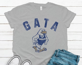 May include: A gray t-shirt with a vintage-style graphic of an eagle wearing a wrestling singlet with the text "GATA" above and "EAGLES" on the singlet.