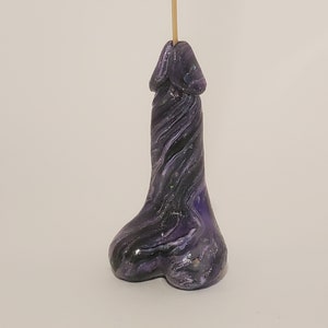 Puede incluir: Porta incienso de color morado y negro con un patrón de espiral y purpurina. El porta incienso tiene forma de parte del cuerpo humano.
