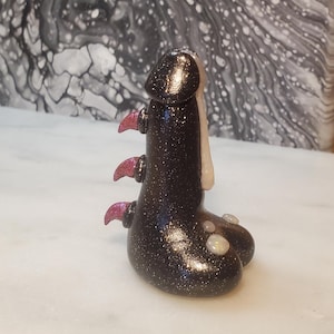 May include: A black and white glittery figurine with pink accents. The figurine has a rounded head with a white detail and three pink spikes on the back.