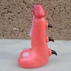 May include: A pink glass figurine with a textured surface and three black claws. The figurine has a rounded shape and a white spot on the bottom.