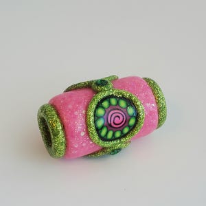 Könnte beinhalten: Eine rosa und grüne Perle aus Polymer Clay mit einem Spiralmuster und Glitzer. Die Perle ist etwa 2,5 cm lang.