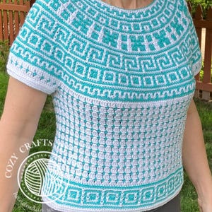 May include: A short-sleeved crochet top featuring a white and teal geometric pattern around the neckline and hem. The main body consists of a textured white stitch pattern. A circular logo with the text COZY CRAFTS is visible on the lower left.