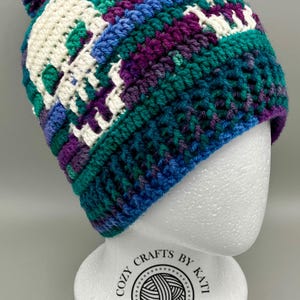 May include: A crocheted beanie featuring a multi-colored pattern of teal, purple, and blue yarn with white skull motifs. The hat is topped with a matching pom-pom and has a ribbed brim. The design incorporates a repeating skull graphic.