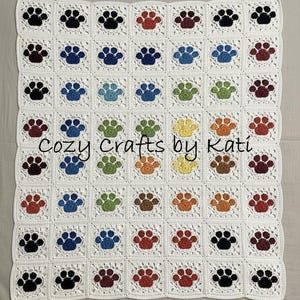 Puede incluir: Una manta de ganchillo con un patrón repetido de coloridas huellas de patas en cuadrados. Las huellas de patas son de varios colores, incluyendo negro, azul, verde, naranja y marrón, sobre un fondo blanco. El texto "Cozy Crafts by Kati" es visible.