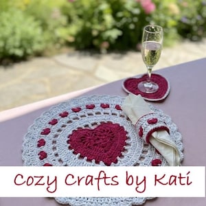May include: A crocheted placemat, coaster, and napkin ring set with a heart design. The placemat is white with a red heart in the center and red heart accents. A glass of champagne sits on the coaster. The text "Cozy Crafts by Kati" is visible.