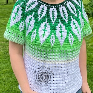 May include: A green, white, and light green crochet top with a leaf pattern design. The top has short sleeves and a round neckline. The bottom of the top is white with a circular logo that says "Cozy Crafts by Katie".