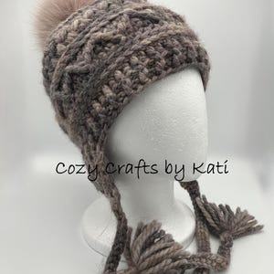 May include: A handmade, crocheted winter hat in shades of gray and brown, topped with a fluffy, light pink pom-pom. The hat features ear flaps with tassels and the text "Cozy Crafts by Kati".