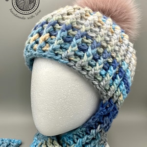 May include: A chunky knit beanie with a variegated pattern of blue, grey, and cream yarn, featuring a large, fluffy, light pink faux fur pom-pom on top. The hat has a thick, ribbed texture and is displayed on a white mannequin head.
