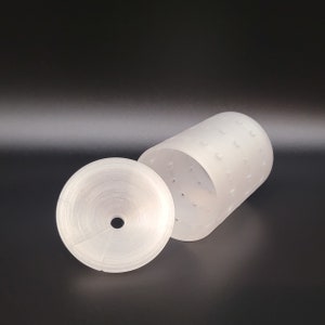 Puede incluir: Un molde de queso de plástico blanco con fondo extraíble. El molde tiene una superficie texturizada y una serie de pequeños agujeros para el drenaje.