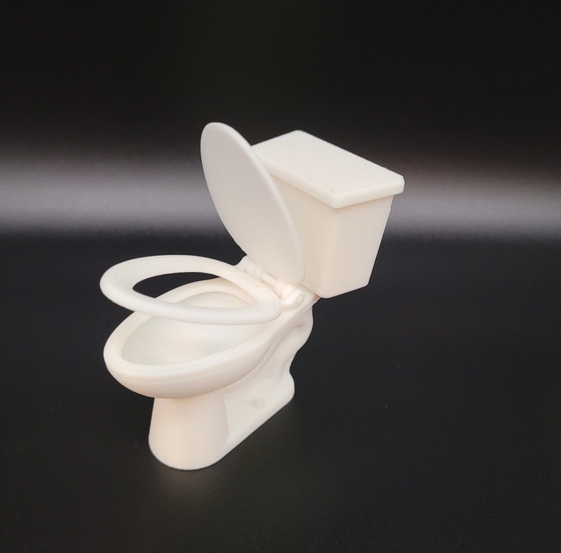 Op de afbeelding: Witte plastic miniatuurtoilet met een gesloten deksel en een aparte zitting. Het toilet is ontworpen voor poppenhuizen of miniatuursettings.