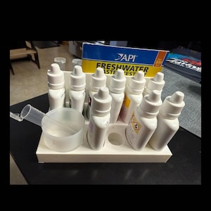 May include: A white plastic organizer holding several white bottles with white caps. A clear plastic scoop and a box labeled "API Freshwater Master Test" are also present. The bottles are arranged in a white plastic holder.
