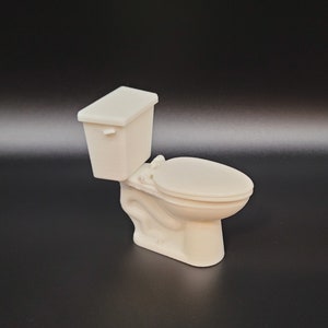 Op de afbeelding: Een wit miniatuurtoilet met een gesloten deksel en een tank. Het toilet is gemaakt van plastic en is ongeveer 10 cm hoog.