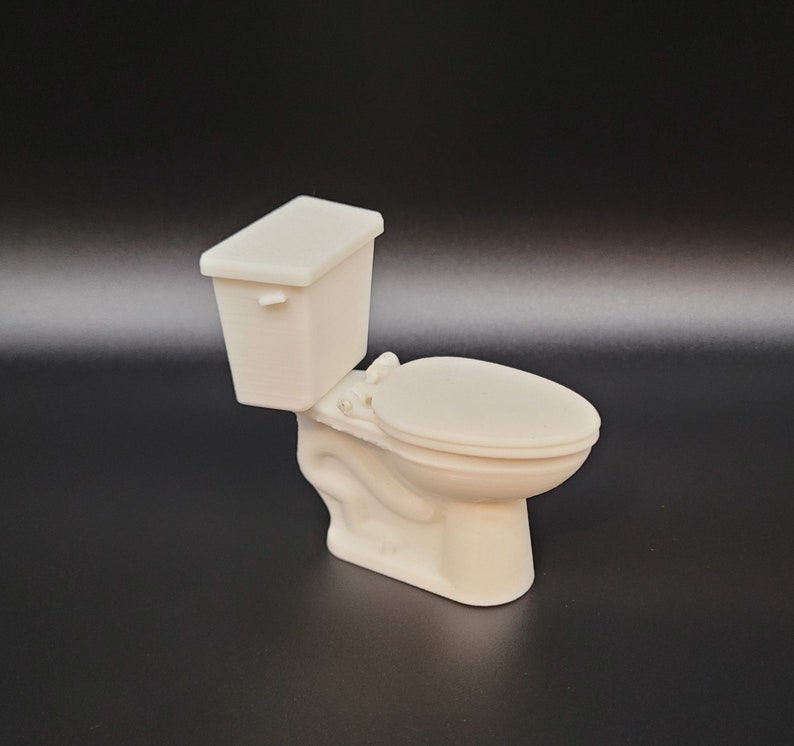 Op de afbeelding: Een wit miniatuurtoilet met een gesloten deksel en een tank. Het toilet is gemaakt van plastic en is ongeveer 10 cm hoog.